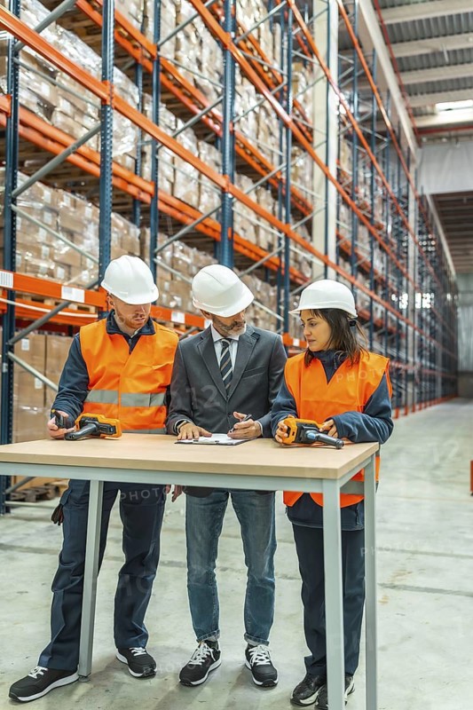 Warehouse workers and manager meeting, discussing logistics and inventory using barcode scanner and clipboard in a large distribution center