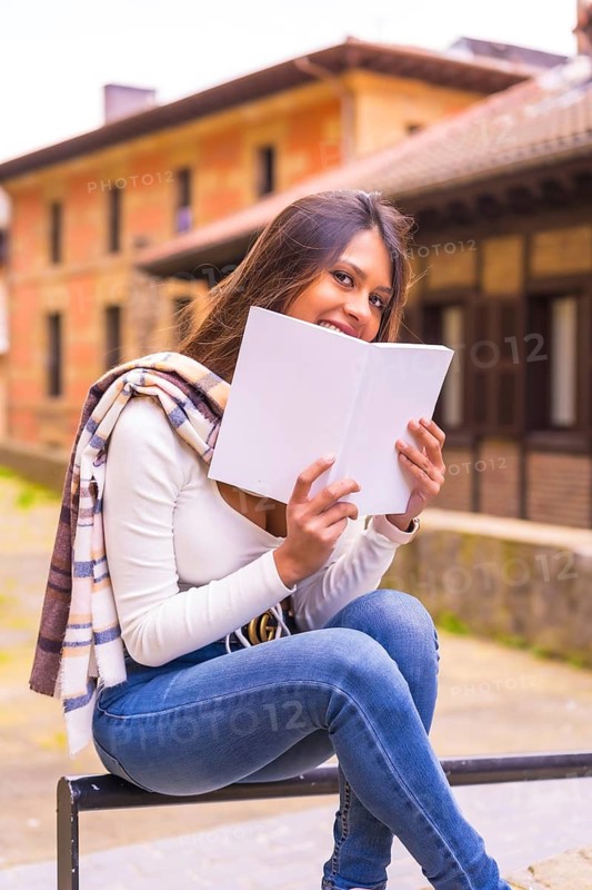 A young Latin woman with a white t-shirt, jeans and a scarf enjoying and imagining reading a book, in the old town. Looking at camera and smiling