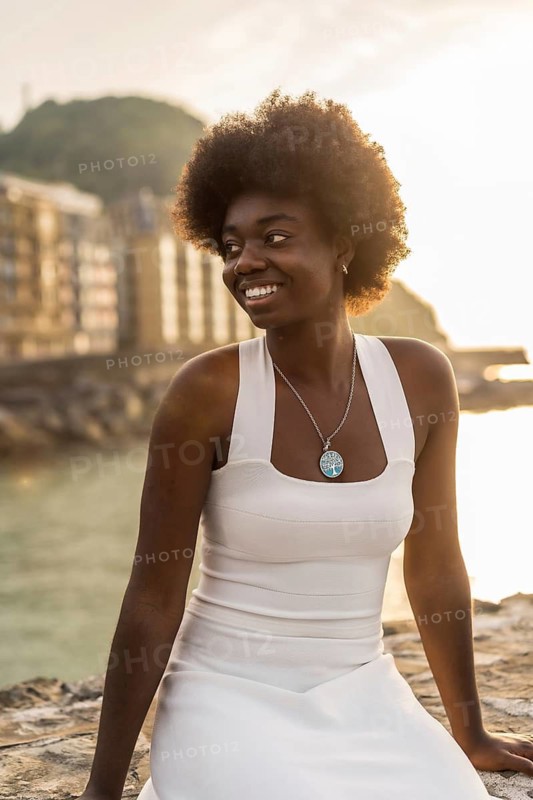 Vertical portrait of a happy beauty african woman in summer white dress sitting outdoors next to the sea during sunset