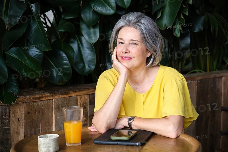 A mature woman with gray hair, wearing a yellow blouse, sits at a wooden table in a cozy setting with a smartphone and a glass of juice, exuding calm and contentment