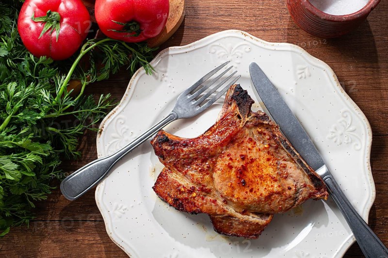 A pork chop rests on a white plate with a fork and knife. Fresh tomatoes and green herbs surround the plate on a wooden table