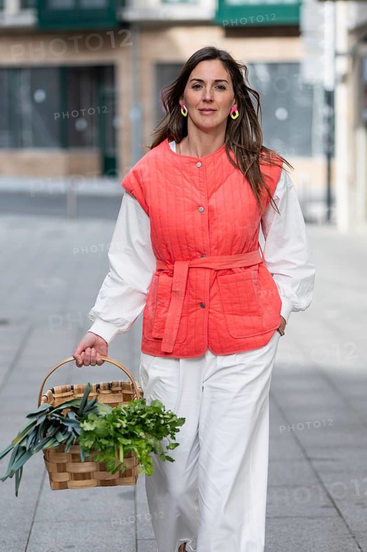 A fashion woman in a red quilted vest and white pants strolls down a city street holding a basket filled with fresh vegetables, exuding effortless style and elegance