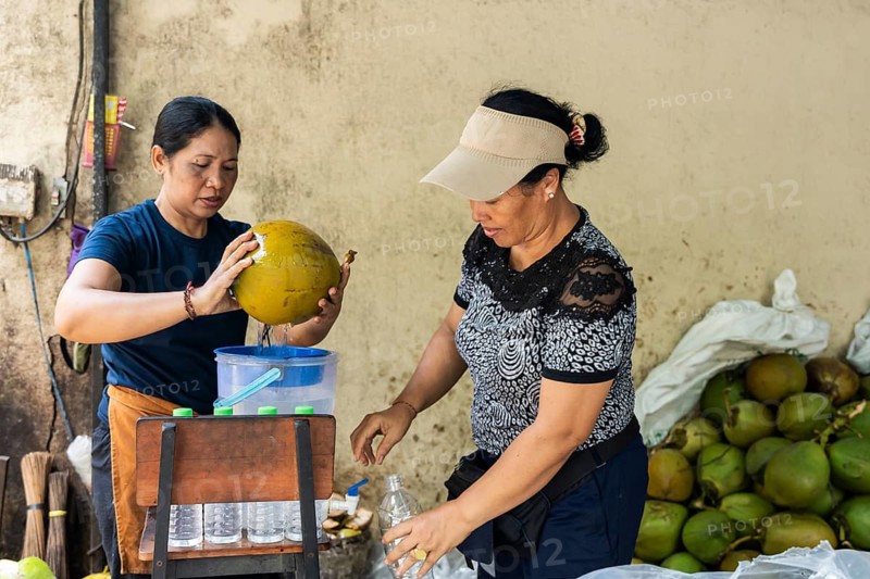 Two asian women expertly open a coconut to extract refreshing coconut water, pouring it into bottles. The setting is a vibrant market with a focus on fresh produce and traditional techniques