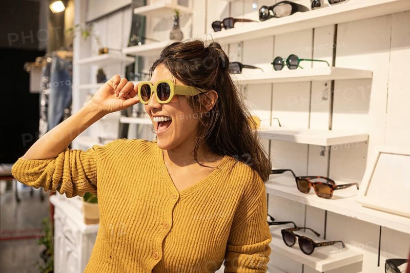 A happy customer tries on a pair of trendy sunglasses in a stylish store, showcasing a wide variety of fashionable eyewear. The bright, inviting setup highlights the fun shopping experience