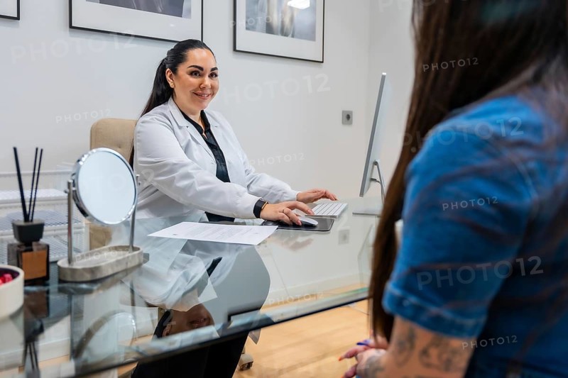 Female doctor in a white coat looking at the camera and smiling, interacting with a patient during a consultation in a modern medical office specializing in aesthetic treatments