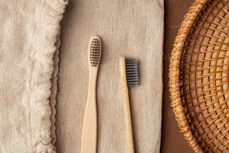 Top view of two bamboo toothbrushes on a textured burlap background, accompanied by a woven basket on a brown surface, showcasing an eco-friendly toiletry setup