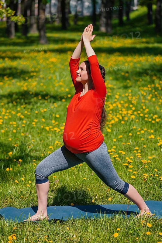 Pregnancy yoga exercise, pregnant woman doing yoga asana Virabhadrasana warrior pose outdoors on grass in summer