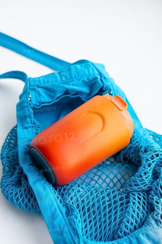 A vibrant orange thermo cup is nestled in a textured blue net bag, portraying a modern, eco-friendly concept against a clean white background