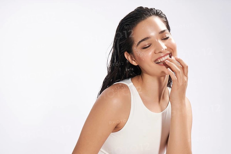 A joyful female model with wet hair and sparkling water droplets on her skin smiles charmingly, caught mid-laugh in a studio setting
