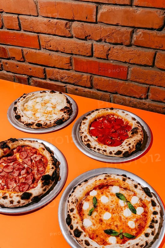 A variety of wood-fired pizzas served on metal plates: Four Cheese with creamy sauce, Margherita with basil, Four Meats with smoked pork, and spicy Pepperoni with chili