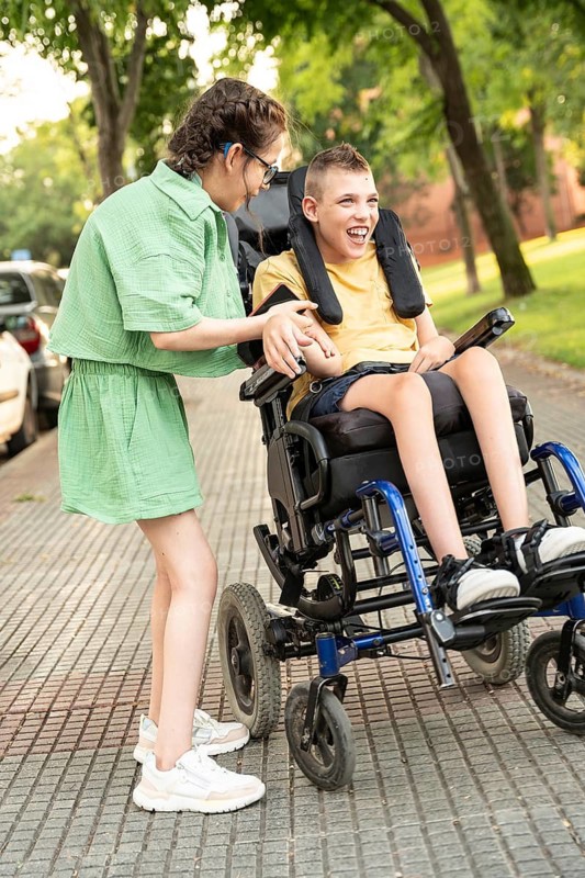 A cheerful girl with a tracheostomy supports her friend with cerebral palsy in a wheelchair, laughing together at the park, surrounded by trees and parked cars