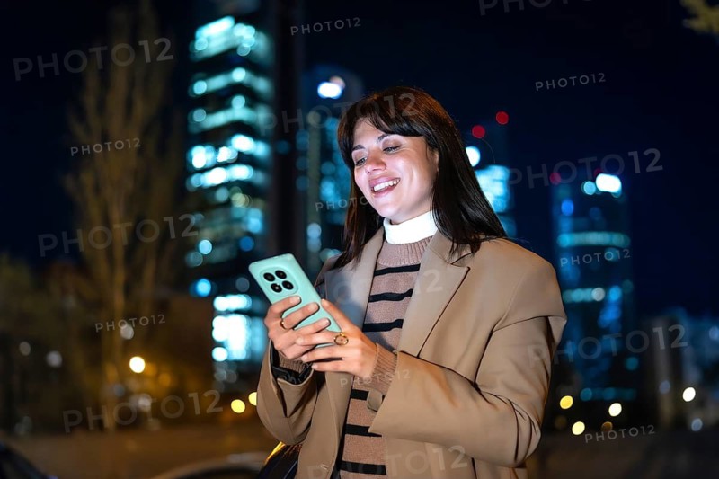 Young woman smiling as she checks her smartphone, screen glow lighting her face against blurred city lights and metropolitan buildings at night, enjoying connected moments