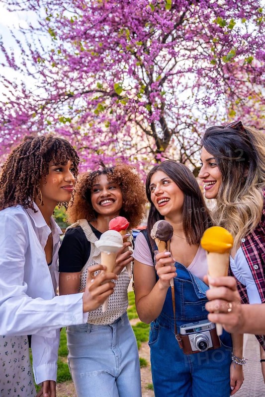 Four female friends are spending time together outdoors, savoring ice cream cones on a sunny spring day under beautiful blossoming trees