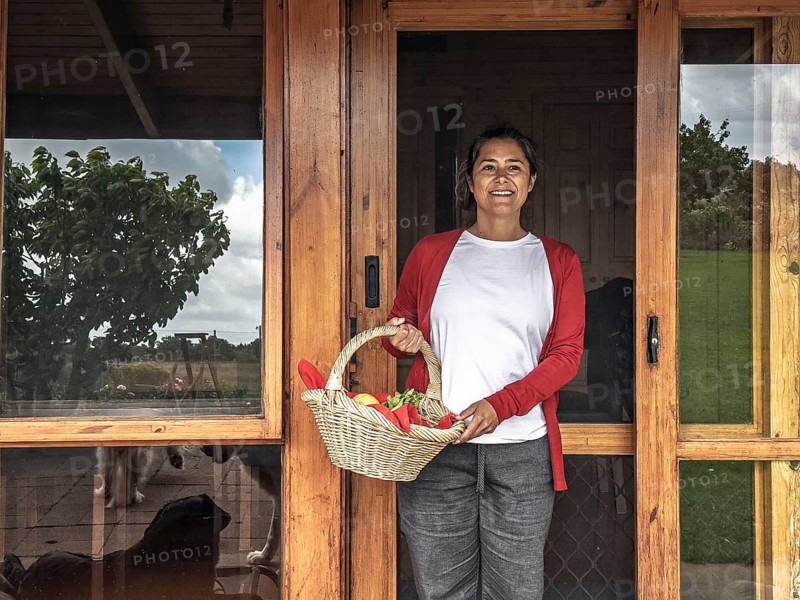 A woman stands at the door of her Australian home, holding a basket of fresh produce, smiling and enjoying her daily routine. The wooden door and outdoor view provide a cozy ambiance