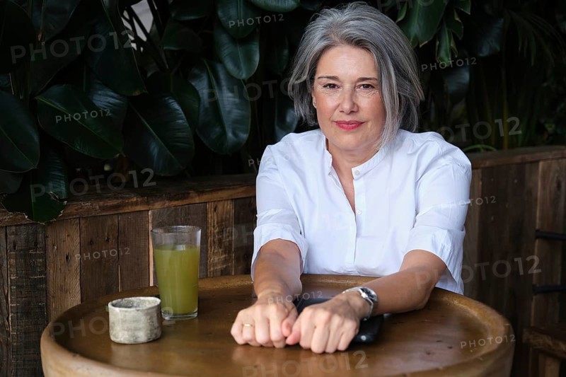 Mature woman sits at a rustic table with a refreshing green drink, surrounded by lush greenery. She appears relaxed and content, embodying a serene and peaceful moment