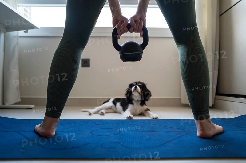 A young woman attempts to exercise at home, holding a kettlebell while her playful dog distracts her on a yoga mat. The warm, cozy setting adds charm to the scene