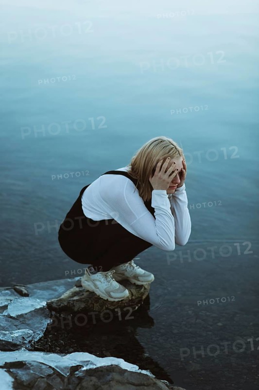 A teenager squats near a calm body of water, with hands covering their face, expressing deep emotion. The tranquil surroundings contrast with the mood depicted