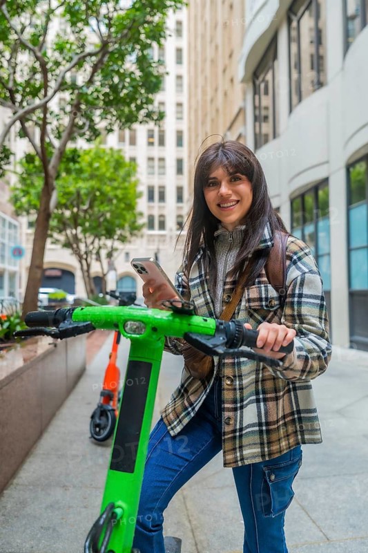 Young woman smiling while holding an electric scooter, checking her smartphone to locate or unlock the shared mobility device on a city street in san francisco