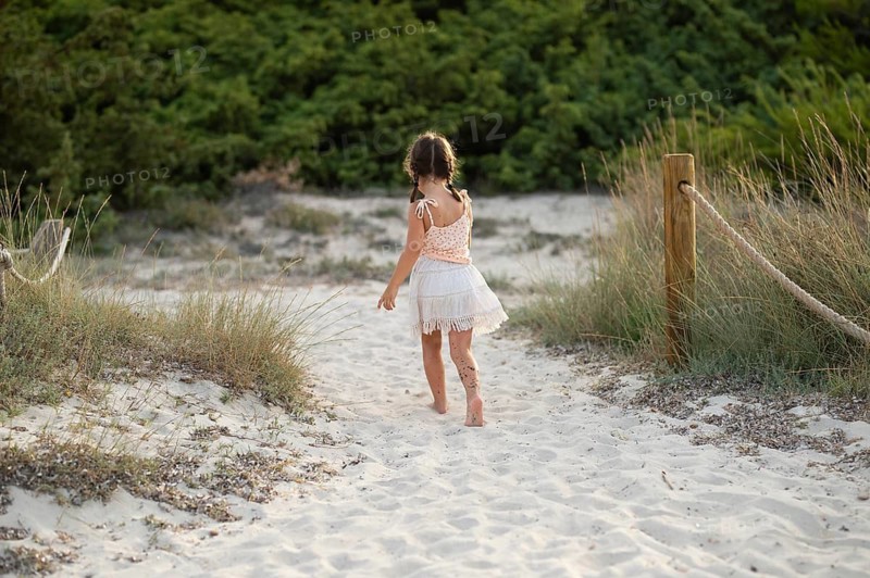 Back view of unrecognizable young girl walking alone on a sandy path by the beach, surrounded by greenery and sandy dunes