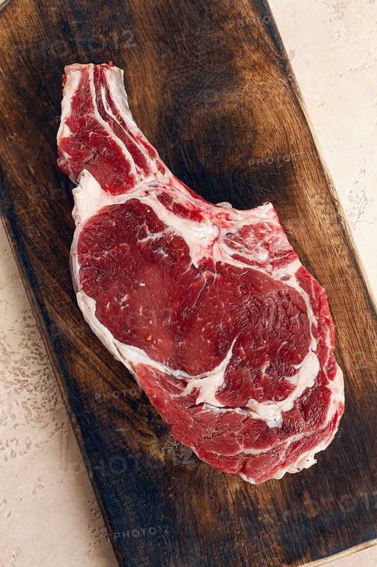 Raw rib eye steak, cowboy steak, wooden chopping board, marble table, top view, no people