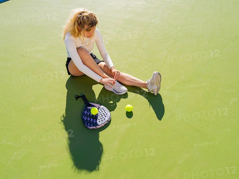 A young woman, captured from above, is tying her shoelaces on a sunlit tennis court, beside her racket and ball