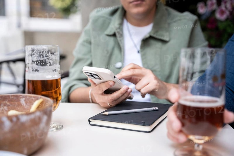 Lesbian couple at a bar chatting while drinking beers. A woman uses a smartphone next to a notebook and pencil, emphasizing connectivity and leisure