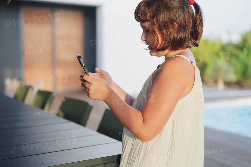 A young girl in a light summer dress stands by the pool, engrossed in her smartphone. The sunny backdrop and serene atmosphere highlight a relaxing summer day