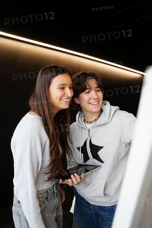 Two teenagers, one non-binary with short hair and one female with long hair, share a joyful moment using a computer in a school environment, highlighting friendship and technology in education