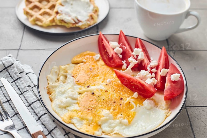 A wholesome homemade meal featuring a fried egg with feta cheese and tomatoes paired with Belgian waffles topped with cream cheese, accompanied by a warm cup of coffee