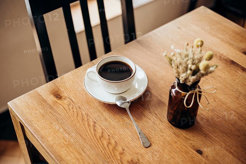 A cozy scene featuring a coffee cup on a wooden table by a window in a cafe. A small vase with dried flowers adds a touch of rustic charm, perfect for a calm morning moment