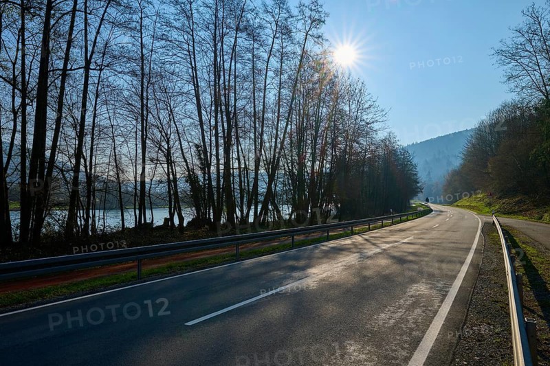 An empty country road lined with trees, with rolling hills in the background and the sun shining through the branches, Faulbach, Main, Spessart, Bavaria, Germany