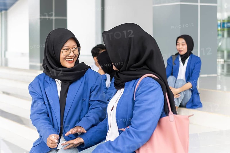 A group of Asian Muslim female students in blue jackets sit together outside a modern school building, engaging in conversation and laughter, bringing a joyful atmosphere to their return to education