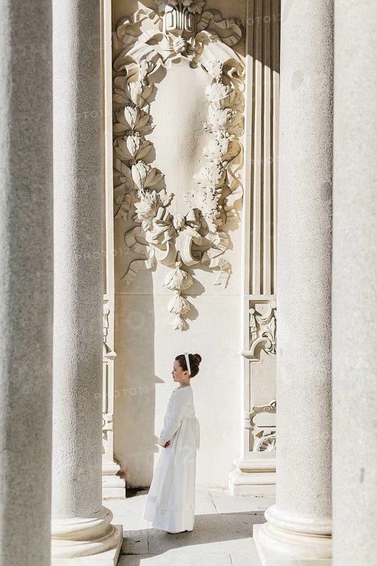 A young girl in a First Communion dress stands gracefully among grand architectural columns and ornate decorations, showcasing elegance and tradition in a serene setting