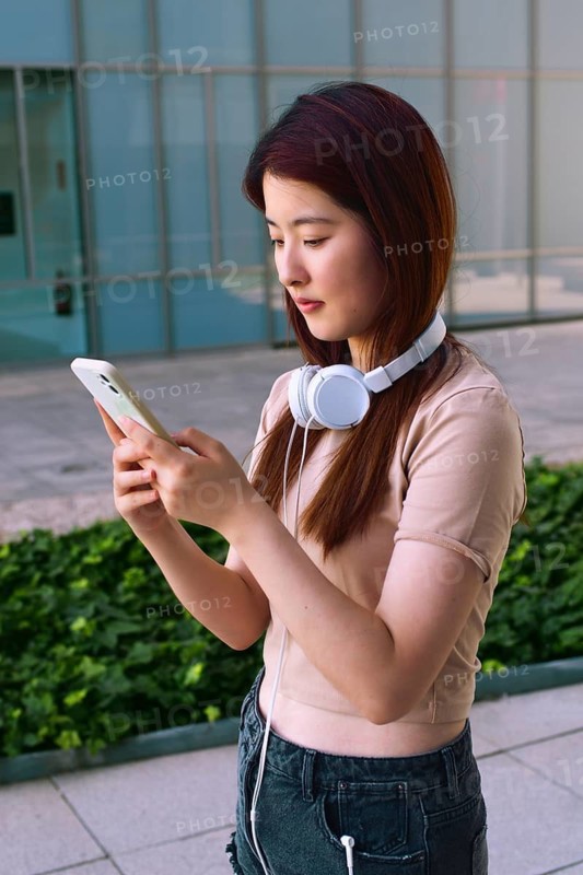 A young woman with headphones around her neck is focused on her smartphone. She stands outdoors with a modern building and greenery in the background
