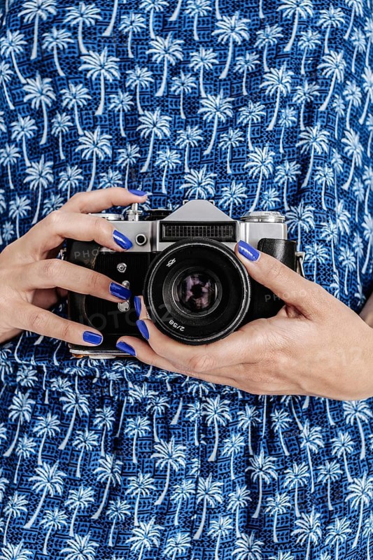 A woman in a blue dress with palm tree patterns is holding a vintage camera. Her fingernails are painted blue, matching the dress, creating a cohesive aesthetic