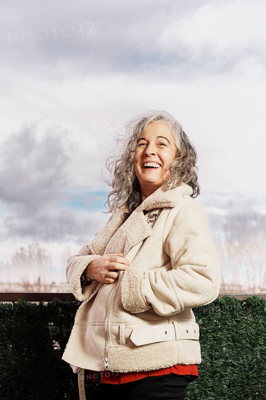 A joyful woman with white hair and a warm jacket stands on her balcony, smiling confidently at the sky, conveying contentment and warmth in a peaceful outdoor setting