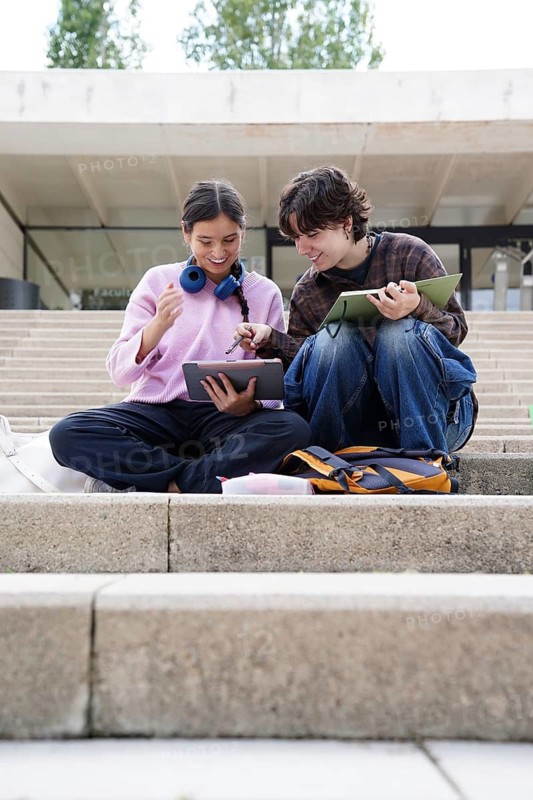 Two cheerful teenagers, a non-binary individual and a female, sit on university steps immersed in their studies. They are engaged in a discussion with a digital tablet and notebooks, symbolizing friendship and learning