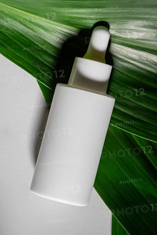 A white cosmetic dropper bottle rests on a vibrant green leaf, highlighting natural and sustainable beauty products in a fresh, minimalist setting