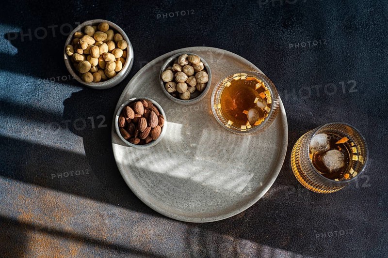 Top view of a rich spiced cognac cocktail accompanied by cinnamon, anise star, and assortments of nuts including almonds, peanuts, and hazelnuts, elegantly presented on a textured tray