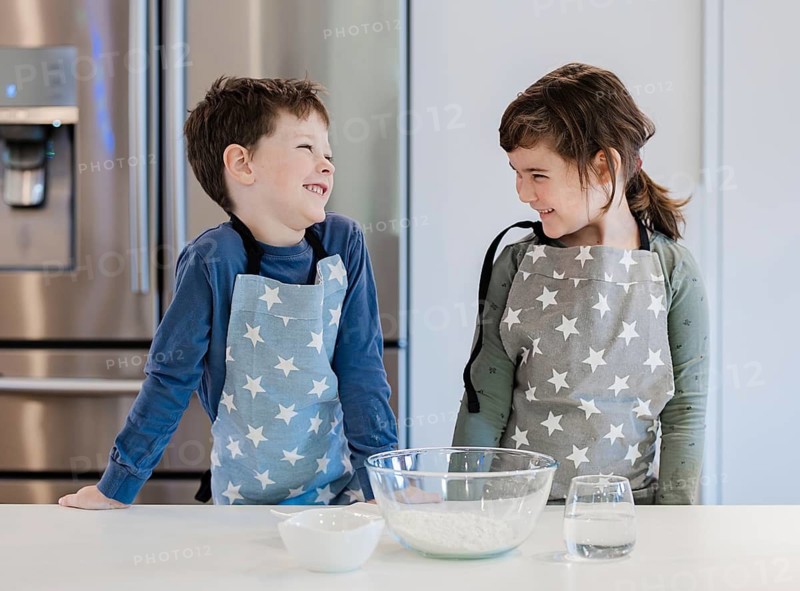 Two siblings wearing star patterned aprons smile at each other in a kitchen. A mixing bowl with flour and a cup of water are on the counter, creating a warm, playful atmosphere