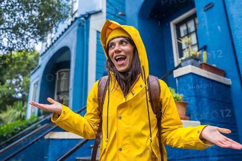 Woman in a yellow raincoat standing happily outside a vibrant blue house, smiling broadly and embracing the rainy weather with open hands during an urban adventure in san francisco