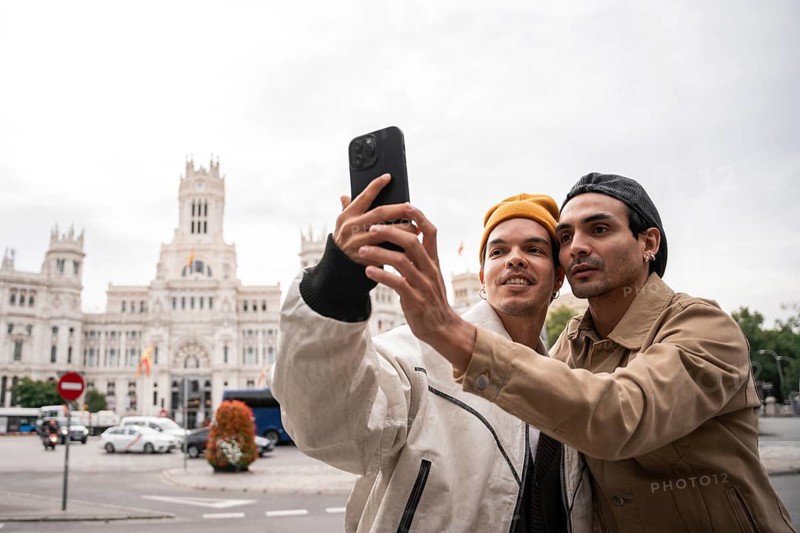A gay couple takes a selfie in front of a historic Madrid building. They are dressed warmly and capture the moment on a smartphone. The street is busy with traffic