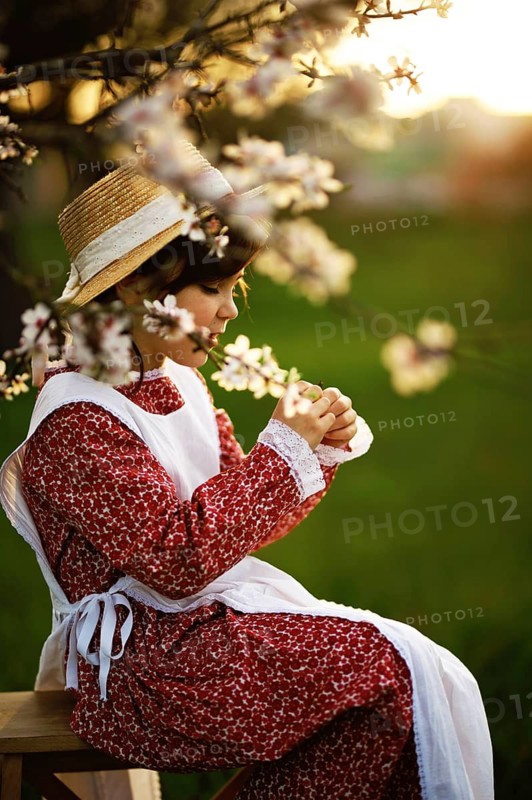 A young girl in a straw hat and vintage floral dress sits on a bench, surrounded by blooming branches in a serene spring setting bathed in warm sunlight
