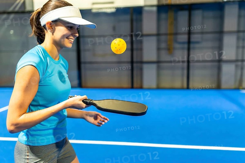 Female athlete smiling while playing pickleball on a blue indoor court, holding a paddle as the ball hovers midair, showing fitness, agility, and energetic recreational play