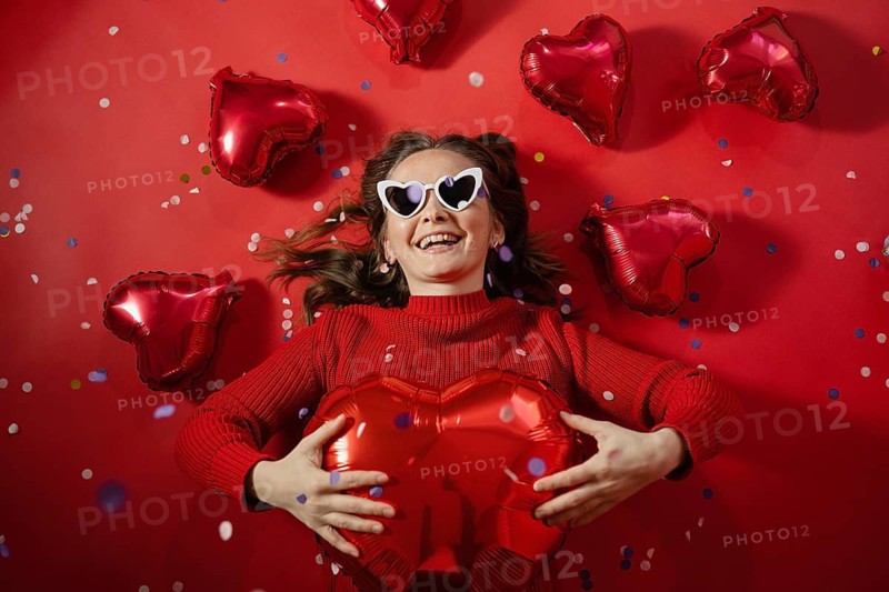 A joyful young woman lies on a red background surrounded by red heart-shaped balloons, holding a large heart balloon on Valentine's day