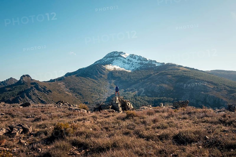 Back view of an unrecognizable person standing on a rock at the summit of Mount Ocejon, in Guadalajara's Sierra de Ayllon, looking towards the snowy peak
