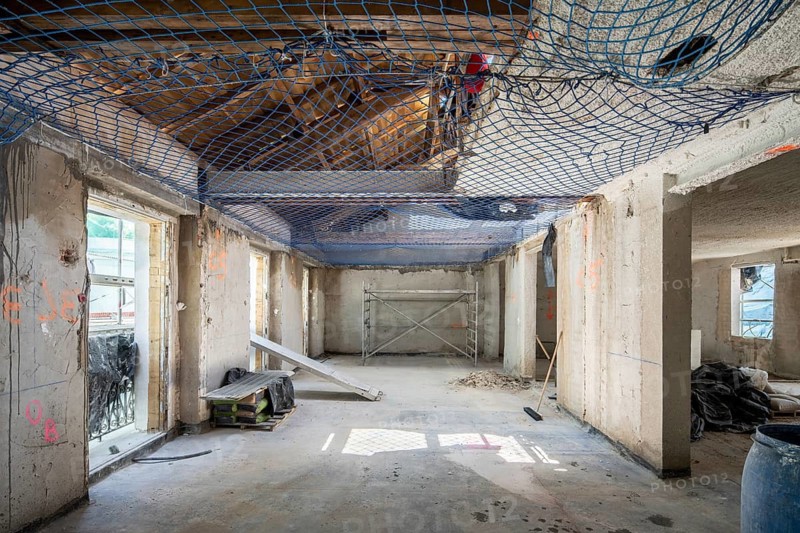 An indoor construction site with exposed concrete walls, safety netting, scaffolding, and construction materials. Sunlight streams through large windows, casting shadows