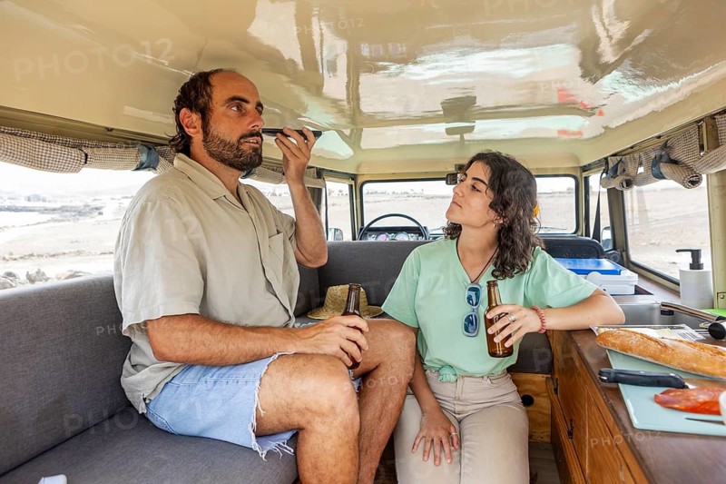 A blind man uses a mobile phone while holding a drink, seated inside a motorhome with a woman. She also holds a drink as they enjoy a relaxed moment during their adventure