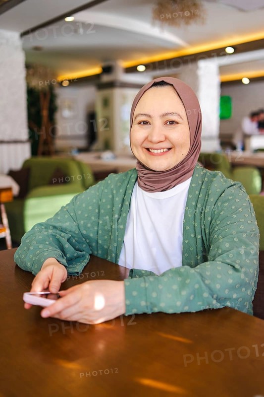 A smiling Muslim woman in a hijab sits comfortably at a table indoors, holding a smartphone. She exudes warmth and positivity, highlighting cultural diversity and modernity