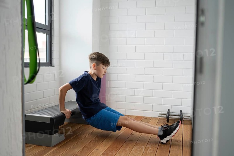 Young boy performs a seated dip exercise on a fitness bench indoors. The room has a modern design with white brick walls, wooden floors, and natural light from a window